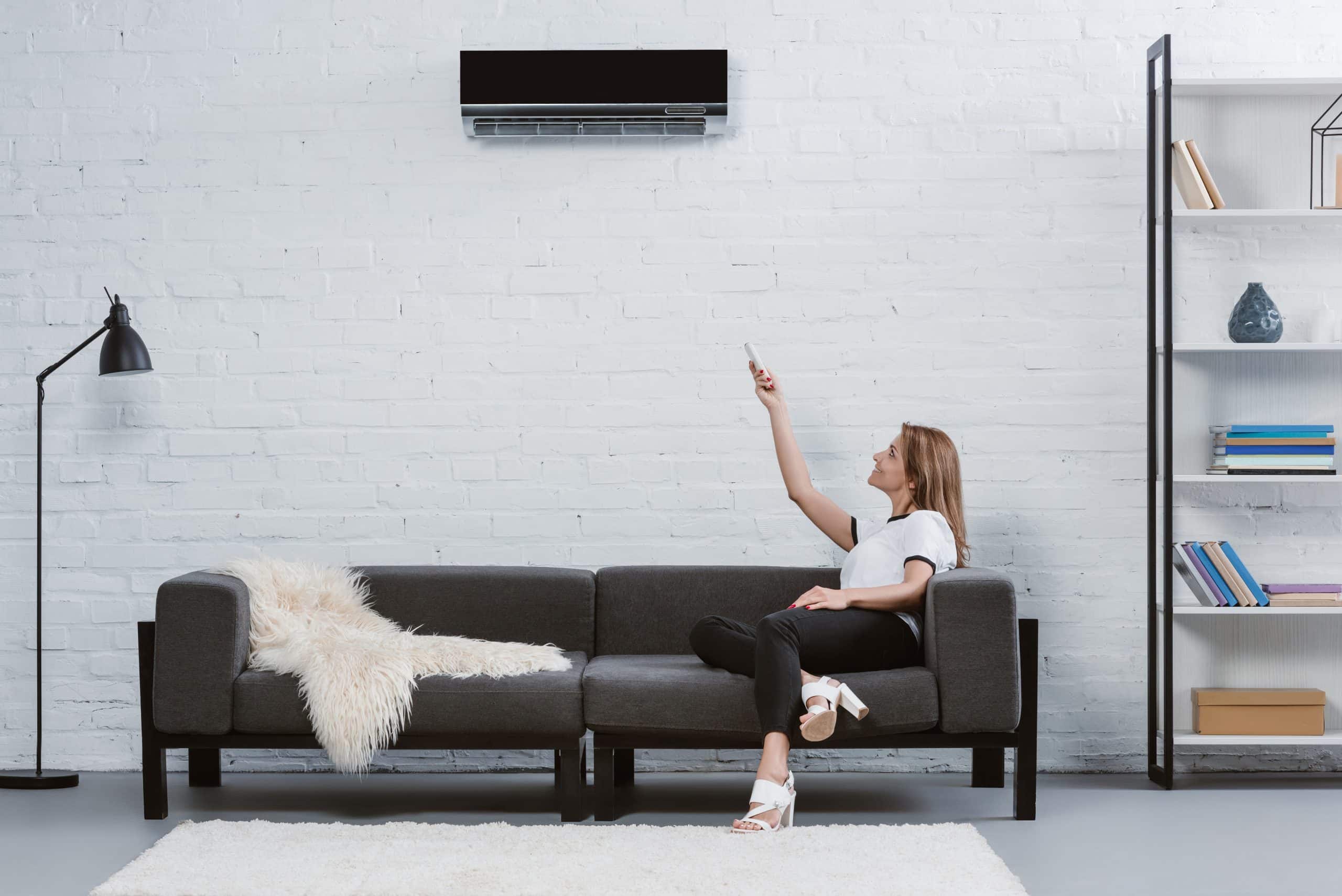 beautiful young woman sitting on sofa and pointing at air conditioner hanging on wall with remote